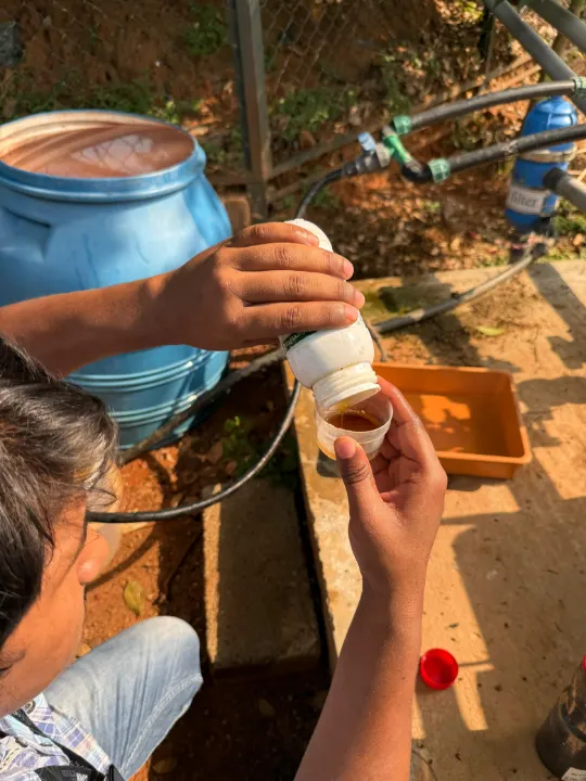 Hands pouring liquid from a bottle into a measuring cup beside drip pipes